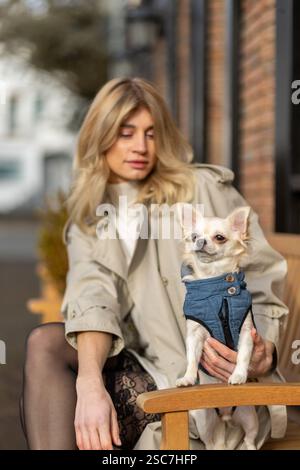 A woman relaxes on a wooden bench in a cozy outdoor setting, holding a small dog dressed in a denim jacket, enjoying the warm sunlight and fresh air. Stock Photo