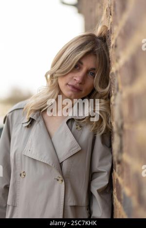 A woman stands confidently with her head against a textured brick wall, wearing a stylish trench coat and displaying a serene expression in a cityscap Stock Photo