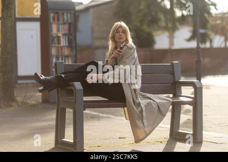 A woman sits comfortably on a bench in a park, enjoying a sunny day while dressed in a fashionable trench coat and black pants. The background feature Stock Photo