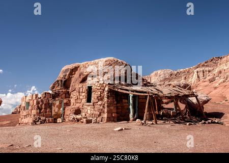 A small house made of stone sits in the desert. The house is covered in a thick layer of dirt and has a wooden porch Stock Photo