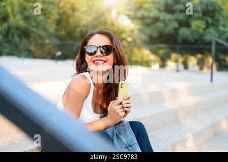 Cheerful young Caucasian woman sitting on a stairway in the street holding a mobile phone Stock Photo