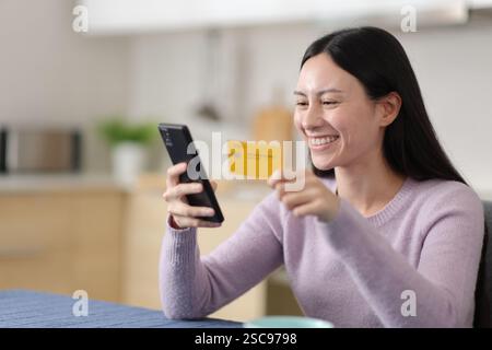 Happy asian woman buying online using card and phone in the kitchen at home Stock Photo