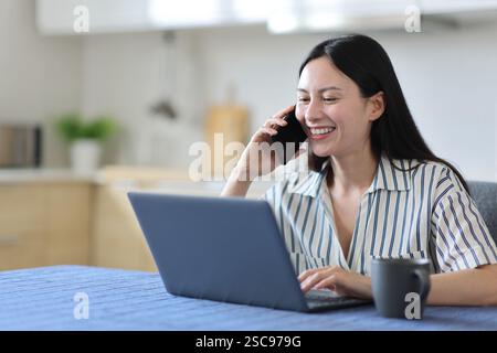 Happy asian woman talking on phone checking laptop in the kitchen at home Stock Photo
