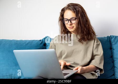 Caucasian woman wearing glasses using laptop while sitting on blue sofa at home. Stock Photo