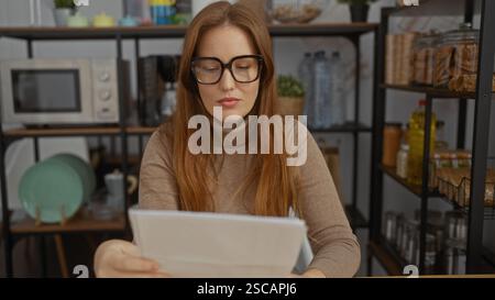 Woman reading document in cozy office setting with glasses and red hair, surrounded by kitchen items and shelves, indicating a casual work environment Stock Photo