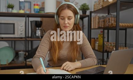 Woman working indoors writing notes with headphones at desk in office setting focused and concentrating. Stock Photo