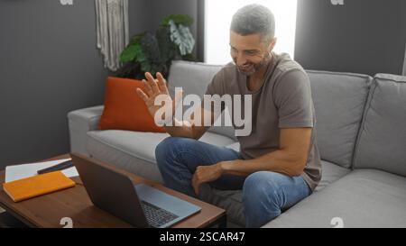 Young man working remotely at home using laptop while sitting on comfortable sofa in cozy living room, displaying a friendly gesture and engaging in v Stock Photo