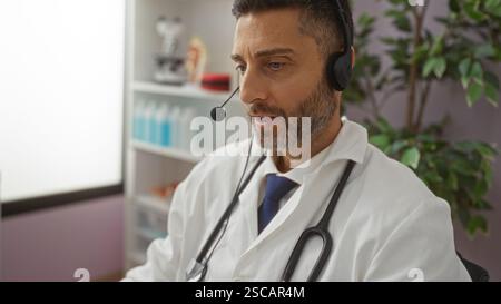 Male doctor using headset in hospital office with medical tools and plants in background, providing virtual consultation services, dressed in white co Stock Photo