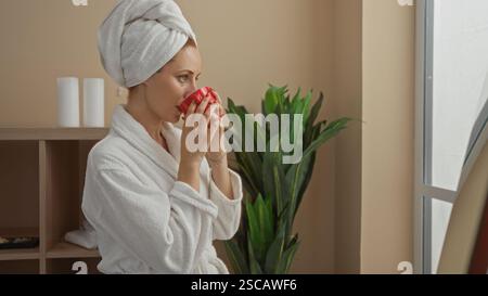 Woman relaxing in a wellness spa center, wearing a bathrobe and towel on her head, drinking tea from a red cup, standing by the window in a serene ind Stock Photo