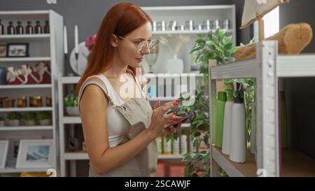 Woman shopping in a decorated store wearing glasses and apron while using a smartphone surrounded by home decor items and cosmetics Stock Photo