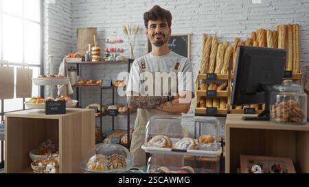 Young man with crossed arms standing in a bakery shop surrounded by various breads and pastries, posing confidently in an indoor setting. Stock Photo