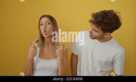 Woman making funny face gestures next to man smiling warmly both wearing white shirts in front of yellow background Stock Photo