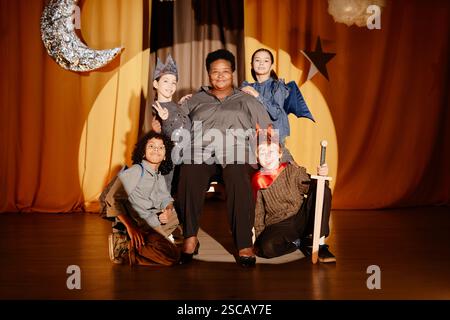 Smiling teacher sitting on stage with diverse group of students wearing costumes, smiling and relaxing after school play event in dramatic setting Stock Photo