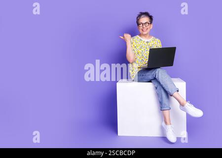 Mature woman smiling and using laptop against purple background in colorful casual attire Stock Photo