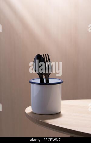 Modern still life: Black plastic spoon and fork elegantly placed in a zinc glass and white enamelware cup, resting on a light wood table. Stock Photo