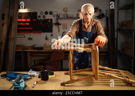 Elderly man restoring wooden furniture in workshop, wearing gloves and apron, surrounded by tools and equipment, focused on craftsmanship and Stock Photo