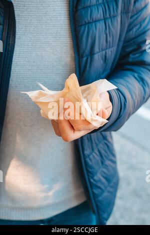 a man, wearing jeans, a gray sweater and a blue jacket, is about to use a recycled paper tissue while is standing outdoors Stock Photo