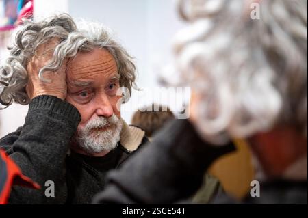 An elderly man with curly gray hair and a thoughtful expression looks at his reflection in a mirror, his hand resting on his head in contemplation. Stock Photo
