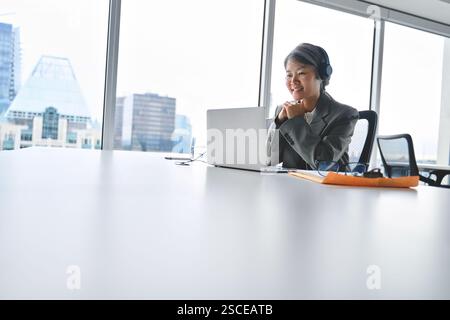 Busy happy mature Asian business woman in headphones using laptop. Stock Photo
