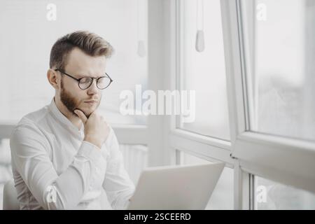 Person with glasses and white shirt concentrating while using a laptop near a window Stock Photo
