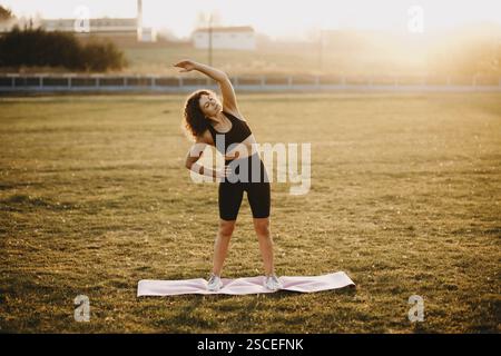Woman performing a side stretch on a yoga mat in the open field Stock Photo