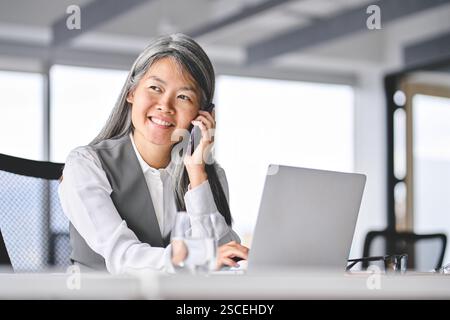 Busy happy older Asian business woman talking on phone making call in office. Stock Photo