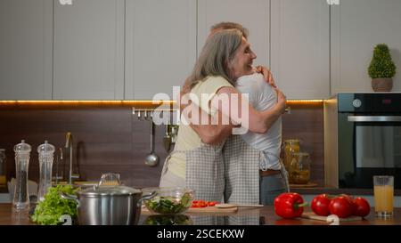 Retired old Caucasian love couple family husband man and wife woman cooking and dancing in kitchen happy grandpa grandma have fun laughing celebrating Stock Photo