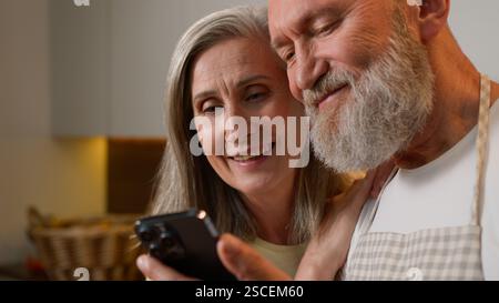 Close up happy old senior Caucasian family laughing in kitchen bonding mature couple grandparents using funny mobile app read news smiling retired man Stock Photo