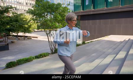 Old Caucasian businesswoman mature woman professional business lady walking up stairs in city outside busy female look at wristwatch late time hurry Stock Photo