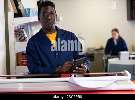 Attentive African American male in a blue uniform using calculator in the printing house Stock Photo