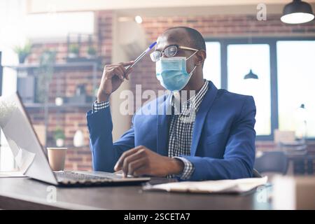 African american man seated at desk, focused on startup analytics with pen in hand. Black male manager in brick wall office tapping his forehead and strategizing new business projects on laptop. Stock Photo