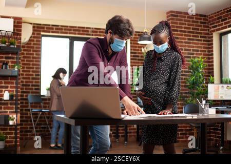 White man and pregnant woman using paperwork and laptop at office desk, planning business project presentation. Diverse colleagues wearing face masks and doing market analysis with documents on table. Stock Photo