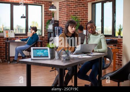 Businesswomen seated together, analyzing articles and sharing ideas during focused discussion using laptop. Tablet displaying company growth statistics as female coworkers collaborate at office desk. Stock Photo