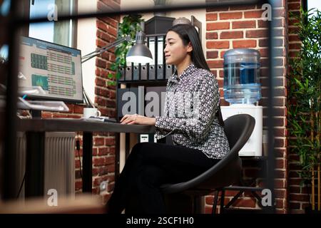 Asian businesswoman examines financial data on her desktop monitor in brick wall workspace. Female entrepreneur analyzes business performance reports on computer, near an office window. Stock Photo