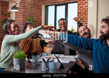 Diverse coworkers joyfully engage in a teamwork activity around desk in a cozy brick wall office. Multiethnic startup group uniting by stacking their hands together after achieving a business deal. Stock Photo