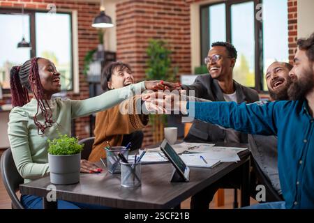 Multiethnic colleagues cheerfully celebrating completion of business project. Diverse startup team seated around table, stacking hands together with excitement, teamwork concept in modern office. Stock Photo