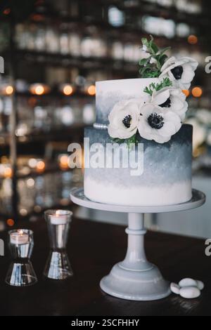 A beautifully crafted two-tier cake in shades of blue is decorated with delicate white flowers. Soft candlelight creates a cozy atmosphere in the back Stock Photo