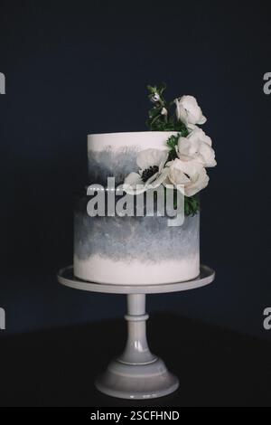 A stunning two-tier cake featuring a gradient gray design adorned with fresh white flowers and greenery, displayed on a simple cake stand against a da Stock Photo