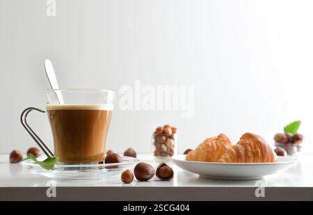 Cup of coffee with hazelnut drink and two croissants on a kitchen bench with fruits around it with white isolated background. Front view. Stock Photo
