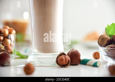 Detail of glass with hazelnut drink with nuts and bowls full of nuts around on white table and green straw with isolated background. Front view. Stock Photo