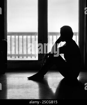 Black and White image of a lonely woman sitting on the floor by the glass door, looking outside. Stock Photo