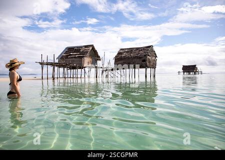 Woman in black swimsuit and a straw hat stadning in water, looking at traditional wooden houses of Bajau people on remote part of Sulawesi, Indonesia Stock Photo