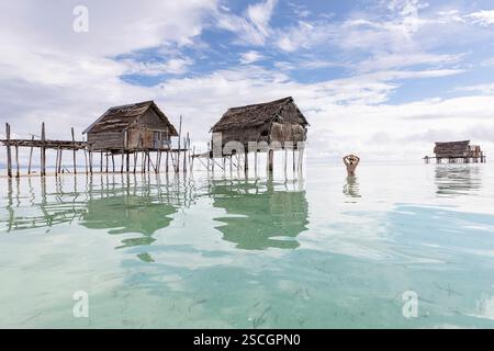 Woman in black swimsuit and a straw hat stadning in water, looking at traditional wooden houses of Bajau people on remote part of Sulawesi, Indonesia Stock Photo