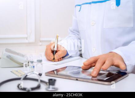 A healthcare worker takes notes and reviews patient data on a tablet while surrounded by medical equipment in a bright office. Stock Photo