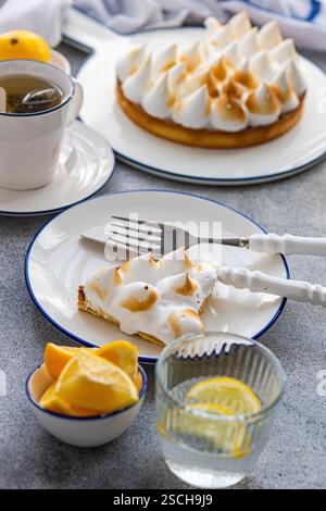 A slice of homemade lemon cake with golden peaks served on a plate, accompanied by a cup of tea, fresh lemon slices, and a lemon garnish in a glass. Stock Photo