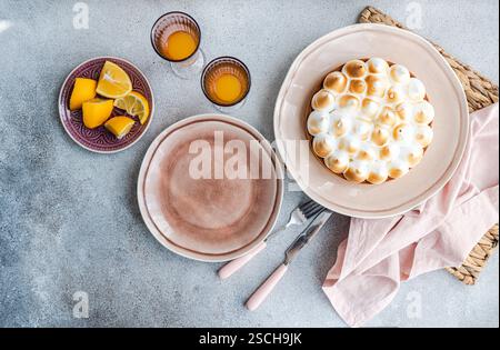 A rustic table setting features a homemade lemon cake with citrus slices on a plate. The arrangement includes empty plates, two glasses filled with or Stock Photo