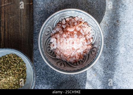 Close-up of Himalayan pink salt in a decorative ceramic bowl alongside fresh green herbs on a rustic wooden and stone background, perfect for healthy Stock Photo