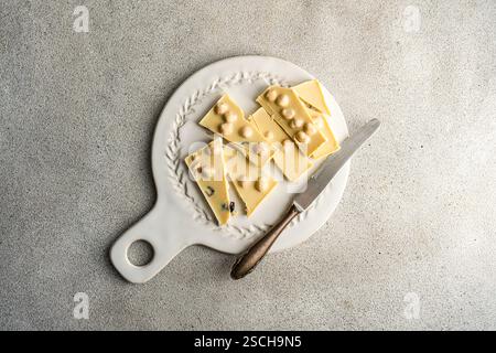 Top view of a white chocolate bar with hazelnuts, broken into pieces on a vintage white plate alongside a rustic knife on a textured grey concrete bac Stock Photo