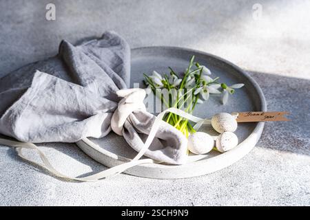 A plate elegantly displays spring snowdrops alongside speckled Easter eggs, a grey linen cloth, and a Thank You tag, all set on a textured surface. Stock Photo