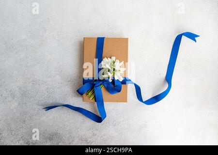 Top view of a gift box wrapped in brown paper, adorned with a vibrant blue ribbon and fresh spring snowdrops placed on a textured grey background. Stock Photo